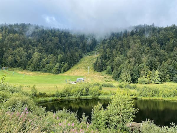 Station de ski en été avec lac au premier plan et verdure avec télésiège au second plan.