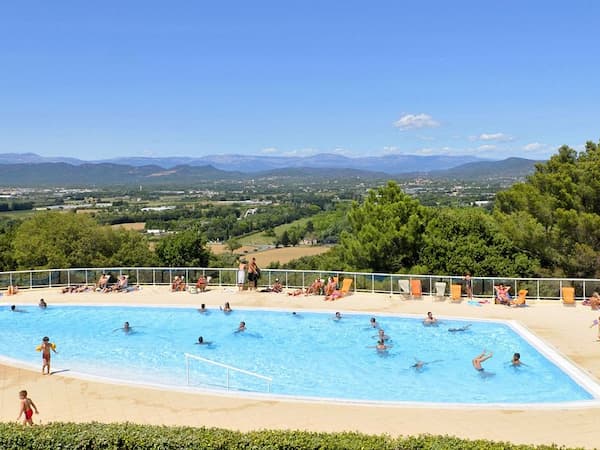 Image d'une piscine au premier plan avec des baigneurs et d'un arrière plan montagneux et forestier.